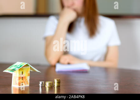 Frau mittleren Alters mit Modell Haus aus Israel Währungen neue israelische Schekel auf hölzernen Tisch. Frau Entspannung zu Hause, Gebäude Haus aus Israel 100 und 50 Banknoten. selektive Fokus auf Geld. Stockfoto