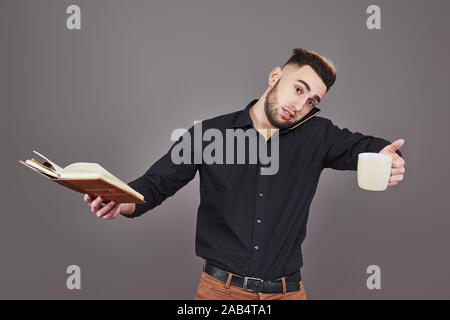 Porträt junger stattlicher Mann mit Telefon, Tasse Kaffee und buchen Sie auf Händen. Der Begriff der Überstunden, Stress und viel Arbeit Stockfoto