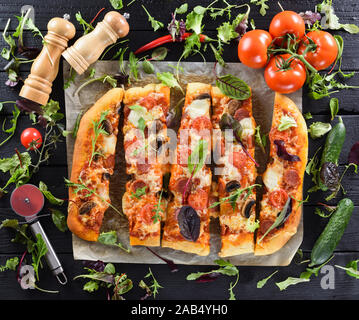 Flatlay von Big leckere Pilze Pizza mit Salami, serviert mit frischen Tomaten, Gurken, Paprika und Kräutern auf schwarzen Hintergrund, Ansicht von oben Stockfoto