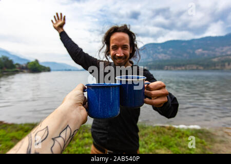 Selektiver Fokus einer tätowierten person Clinchen eine Tasse Kaffee mit einem glücklichen jungen Mann vor einem wunderschönen Natur Landschaft im Freien Stockfoto