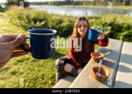 Selektiver Fokus von zwei Menschen jubeln Ihre Tassen Kaffee oder Tee, während Sie ein Picknick an einem sonnigen Tag draußen in der Natur Landschaft Stockfoto