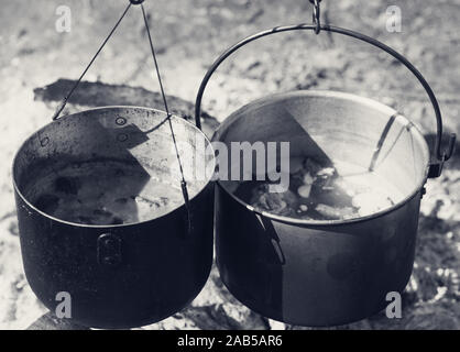 Kochen in zwei alten rußigen Kessel am Lagerfeuer am sonnigen Tag. Schwarze und weiße Farben Landschaft. Stockfoto