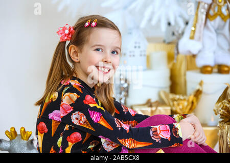 Kleines Mädchen sitzt in der Nähe ein neues Jahr Baum und lächelt. In der Nähe der Box mit einem Geschenk. Stockfoto