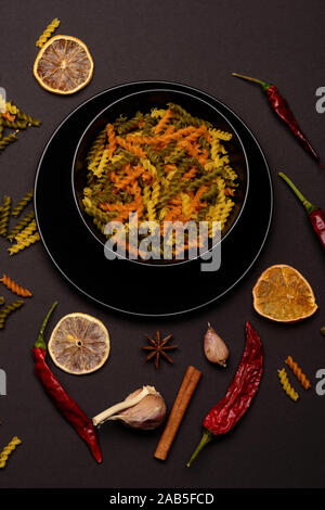 Leckere bunte Italienische Pasta in Keramik schwarz Teller Chili, Orange, Knoblauch, Sternanis und Zimt weiß und blau karierten Stoff auf dunklem Hintergrund, Ansicht von oben Stockfoto