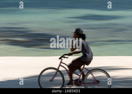 San Pedro, Ambergris Caye, Belize - November, 15, 2019. Ein abstraktes Bild von einen Radfahrer am Telefon zu sprechen in der Bewegung gegen eine blaue karibische Meer b Stockfoto