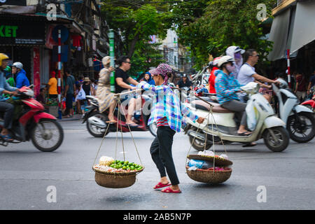 Hanoi, Vietnam - 11. Oktober 2019: asiatische Frau mit einem hölzernen Durchführung pol Transport von Waren über eine befahrene Straße in Hanoi, Asien Stockfoto