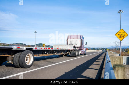 Kraftvoller Big klassische amerikanische Motorhaube rig brown Semi Truck mit hoher Kabine Transport von leeren Flachbett Auflieger, die auf der geraden Straße mit Pro Stockfoto