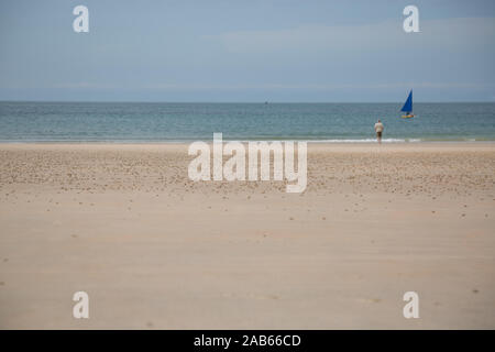 Mann, der am Strand mit Meerblick, mit einem blauen Segelboot vorbei. Jersey, Channel Islands. Stockfoto