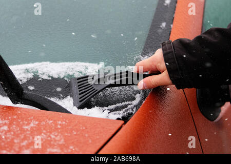 Abstreifer in mans hand. Man löscht Schnee von Vereisten Windschutzscheibe des Autos. Reinigung gefrorenen Fenster von orange Automobil. Snowy. Stockfoto