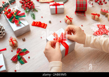 Vorbereitung Geschenke für Weihnachten und das neue Jahr. Handgefertigt. Das Mädchen setzt einen Bogen über das Geschenk. Dekorationen und Geschenke auf hölzernen Tisch. top Aussicht, flach. Stockfoto