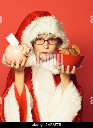 Junge süße santa claus Junge mit Brille in Weihnachten Pullover und neues Jahr Weihnachten hat Holding pink piggy Bank Schwein und Schokolade hip Cookies in Schüssel. Auf Rot studio Hintergrund Stockfoto