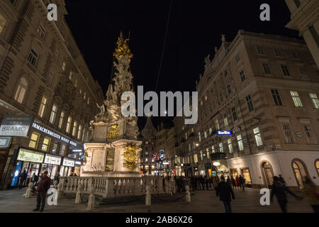 Wien, ÖSTERREICH - NOVEMBER 6, 2019: Wiener Pestsaule, auch genannt die Pestsäule am Graben Straße, in der Nacht, mit Fußgängern vorbei. Es ist ein Stockfoto