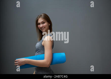 Happy Sportlerin mit blauem Teppich in die Hände auf dem Hintergrund der grauen Wand in der Turnhalle Stockfoto