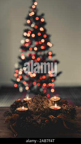 Advent oder Weihnachten Kranz mit vier brennenden Kerzen. Schließen Sie herauf Bild. Unscharf Weihnachtsbaum im Hintergrund. Bokeh der Weihnachtsbeleuchtung. Stockfoto