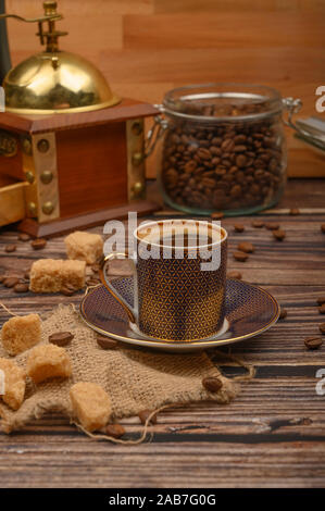 Eine Tasse Kaffee, Kaffeebohnen in einem Glas, brauner Zucker, Kaffeemühle auf einer hölzernen Hintergrund Stockfoto