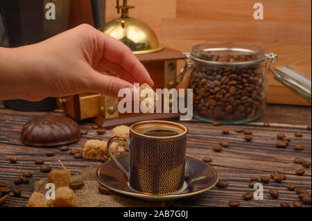 Hand Mädchen setzt ein Stück braunen Zucker in einer Tasse Kaffee, Kaffeebohnen, brauner Zucker, Kaffeemühle auf einer hölzernen Hintergrund Stockfoto