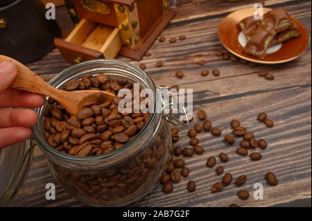 Die Hand des Mädchens nimmt einen Löffel aus Holz von Kaffeebohnen aus Glas, eine Kaffeemühle, Stücke Schokolade auf einem hölzernen Hintergrund. Nahaufnahme Stockfoto