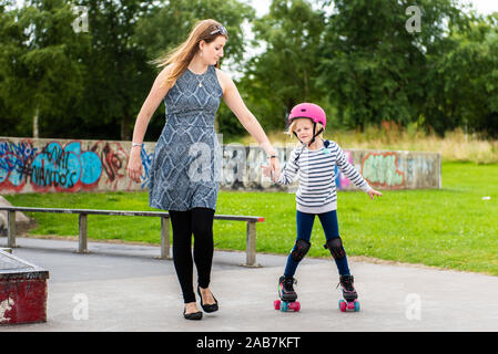 Ein junges Mädchen mit einem rosa Helm hält ihre Mütter Hand, mit der sie zu Roller Skate lernt Am Skatepark, fit, jung und gesund Stockfoto