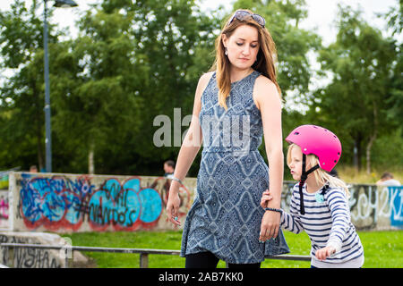 Ein junges Mädchen mit einem rosa Helm hält ihre Mütter Hand, mit der sie zu Roller Skate lernt Am Skatepark, fit, jung und gesund Stockfoto