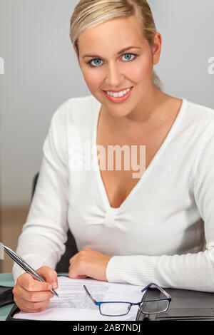 Porträt einer wunderschönen jungen Frau oder Geschäftsfrau mit perfekten weißen Zähnen an ihrem Schreibtisch arbeiten in einem Büro schreiben und lächelnd in die Kamera Stockfoto