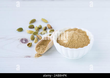 Eine Nahaufnahme von einem kleinen weißen Schale gefüllt mit gemahlener Kardamom mit einer kleinen Schaufel der gleichen und einige ganze Kardamomsamen dahinter. Stockfoto