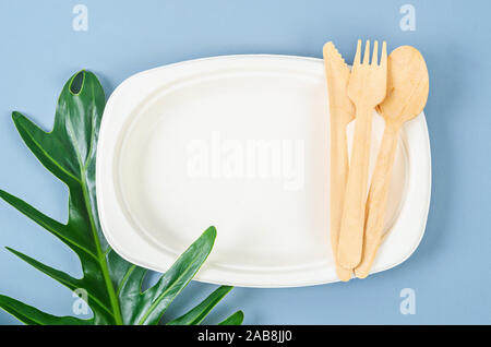 Teller aus Bagasse für Lebensmittel und Löffel aus Holz mit grünen Blättern auf blauem Hintergrund. Umweltfreundliches Konzept. Stockfoto