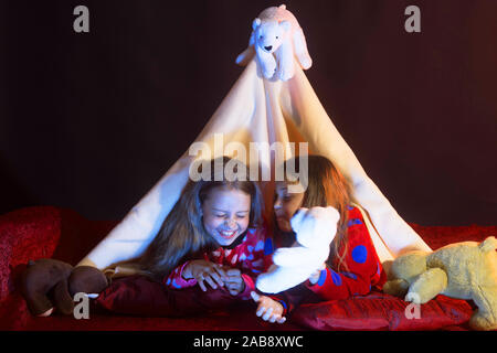 Mädchen mit glücklichen Gesichtern liegen unter Decke Zelt auf dunklem Hintergrund. Kindheit und Glück Konzept. Kinder im Schlafanzug mit Decke spielen mit Weißer Teddybär abgedeckt. Kinder haben Pyjamaparty Stockfoto