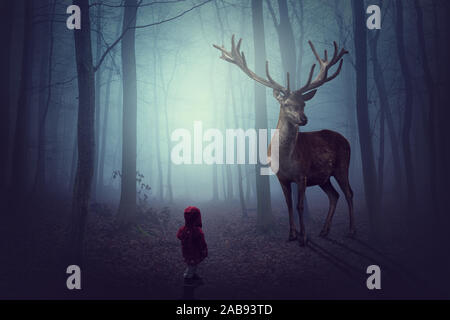 Ein kleines Kind mit roter Jacke steht vor einem großen Hirsch im dunklen Nebelwald Stockfoto