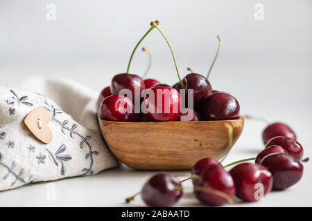 Rote Kirschen auf einem weißen Hintergrund. Stillleben mit Kirschen in eine hölzerne Schüssel und ein beige Serviette mit einem Holz- Knopf Herz Süß. Saftige frische reife che Stockfoto
