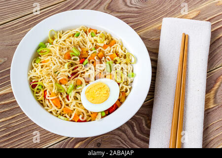 Instant Nudeln mit Ei und Gemüse in weiße Schüssel und Essstäbchen auf Tisch Serviette über eine braune Holztisch. Komfort fast food. Ansicht von oben. Stockfoto