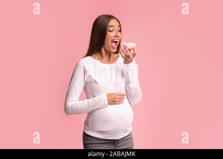 Schwangere Frau beißen Donut steht auf rosa Hintergrund, Studio Stockfoto