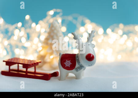 Close-up Keramik Weihnachten Rentier Spielzeug, Fir Tree Kerze und Schlitten auf verschneiten und defokussierten Hintergrund leuchten. Weihnachten oder Silvester feiern. Stockfoto