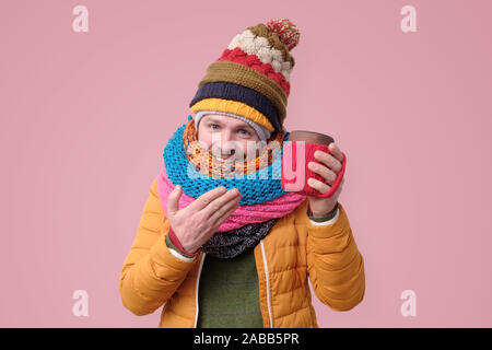 Stattliche hipster Mann in Mütze, Schal Schale hält mit heißem Kaffee oder Tee. Studio shot auf rosa Hintergrund. Stockfoto