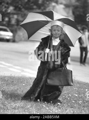 1980 s, historischen, außerhalb Holding ein Regenschirm, ein süßes kleines Mädchen 'modelling' ist die vorherige Generation Dame Bekleidung und Zubehör; einen dicken Pelzmantel, Schnürung, Hut und Handtasche, alle an der Flohmarkt ist eine ihrer Schule, England, UK verkauft zu werden. Stockfoto