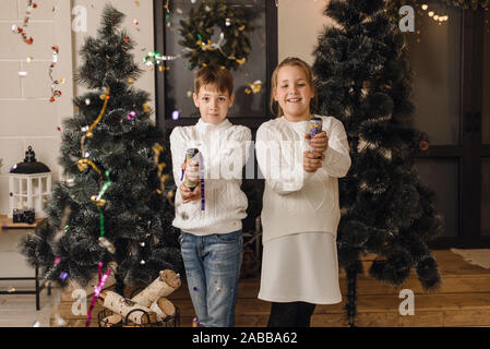 Zwei süße Kinder auf dem Hintergrund der Weihnachtsbäume öffnen Cracker und Spaß haben. Bruder und Schwester in Weiß gestrickte Pullover feiern das neue Jahr und Stockfoto