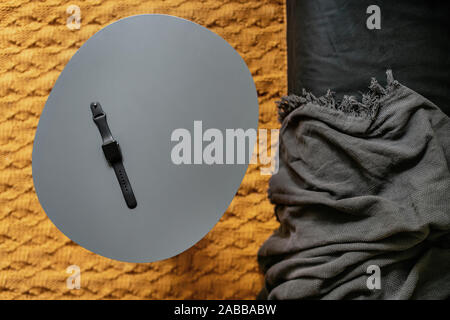 Kleine Metall grau Tabelle mit einem Black Watch und einem dunklen Sofa mit einem Plaid auf der hellen orange Teppich im Innenbereich. Closeup top Foto anzeigen. Horizontale. Stockfoto
