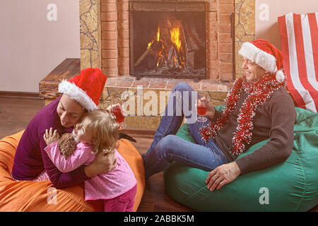 Ein Ehepaar, ein Mann und eine Frau in santa Hüte auf dem Kopf, Sitzen vor dem Kamin in Bohne Beutel mit einem kleinen Kind, eine und eine Hal Stockfoto