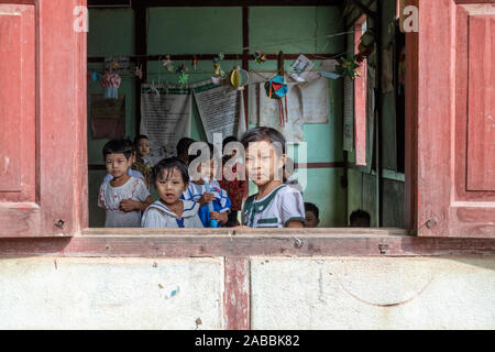 Schüler der Grundstufe lean aus ihrem Klassenzimmer Fenster mit Lächeln und Neugier in der Kanne Dorf am Chindwin Fluss von Myanmar (Birma) Stockfoto