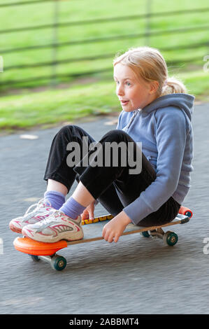 Junge blonde Mädchen sitzt auf einem sich schnell bewegenden Skateboard aufgeregt. Panning Action Shot mit Motion verschwommenen Hintergrund Stockfoto