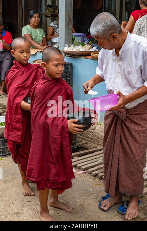 Zwei junge buddhistische Mönche in roten Roben gekleidet geben Sie den Markt der Kanne, Myanmar (Birma) auf der Suche nach Almosen und sind mit einem Reis Spende belohnt Stockfoto