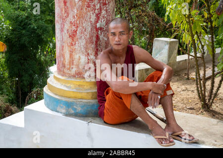 Ein junger buddhistischer Mönch mit rasiertem Kopf in orange und rote Gewand gekleidet liegt von einem pllar seines Klosters entlang der Chindwin Fluss, Myanmar (Birma) Stockfoto