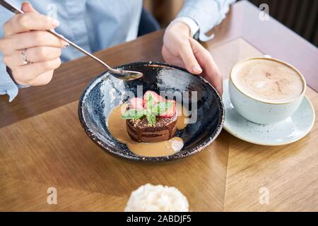 Frau isst Schokolade Biskuit, mit Karamell, mit Erdbeeren und minzeblatt eingerichtet. Frühstück im Café, Kaffee am Morgen. Am Holztisch Stockfoto