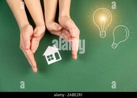 Familie Haus und Immobilien Konzept. In der Nähe von Frau und Kind Hände halten Weißbuch Haus und Glühlampe Bild auf grünem Hintergrund. Flach, Stockfoto