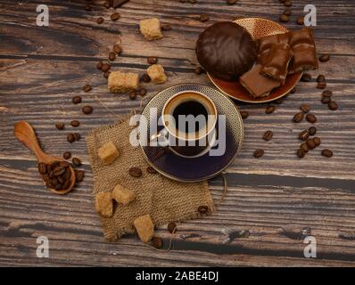 Eine Tasse Kaffee, Kaffeebohnen, brauner Zucker, Schokolade Stücke, Marshmallows in Schokolade auf einem hölzernen Hintergrund Stockfoto