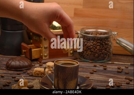 Hand Mädchen setzt ein Stück braunen Zucker in einer Tasse Kaffee, Kaffeebohnen, brauner Zucker, Kaffeemühle auf einer hölzernen Hintergrund Stockfoto