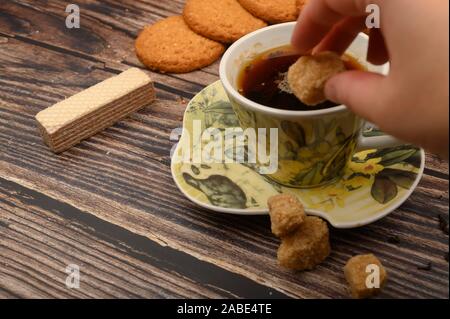 Die Hand des Mädchens legt ein Stück braunen Zucker in einer Tasse schwarzen Tee, oatmeal Cookies, Tee, Waffeln, brauner Zucker auf einer hölzernen Hintergrund. Nahaufnahme Stockfoto