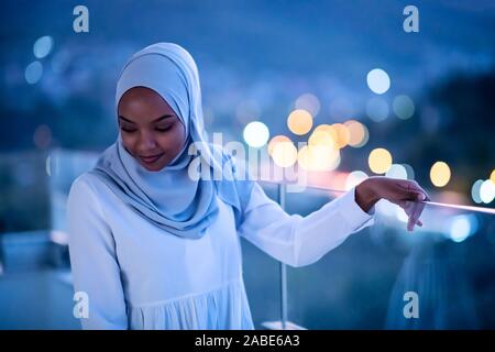 Afrikanische Moderne muslimische Frau in der Nacht am Balkon lächelnd an Kamera mit Stadt bokeh Lichter im Hintergrund Stockfoto