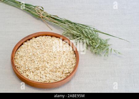 Haferflocken in einer braunen Keramik Schüssel mit auf einem Hintergrund von Grün reif Ohren von Hafer auf Leinen Tischdecke. Das Konzept der Kochen natürliches und gesundes Essen. Stockfoto