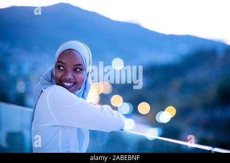 Afrikanische Moderne muslimische Frau in der Nacht am Balkon lächelnd an Kamera mit Stadt bokeh Lichter im Hintergrund Stockfoto