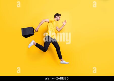 Volle Größe Foto von hübscher Kerl springen hoch halten Pakete Geschwindigkeit hetzen Verkauf shopping Verschleiß casual t-shirt Hose isolierte gelbe Farbe Hintergrund Stockfoto